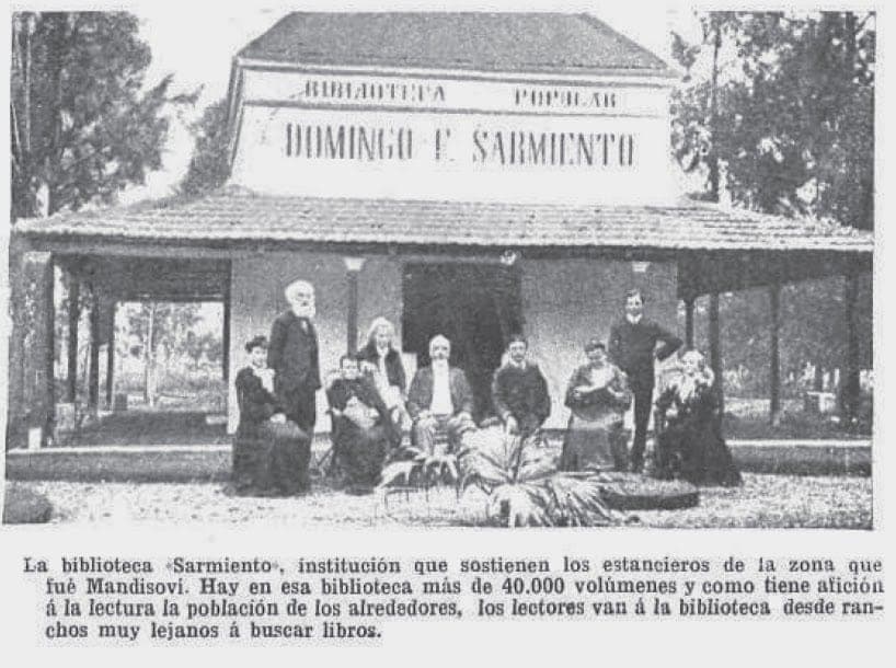 Fotografía de prensa antigua: un grupo de personas posa bajo la galería de un edificio bajo con el cartel «Biblioteca Popular Domingo F. Sarmiento», con su epígrafe impreso debajo.
