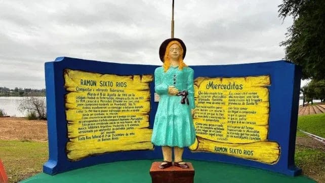 Monumento a Merceditas: una escultura de mujer con vestido celeste frente a un panel azul y amarillo con los textos sobre Ramón Sixto Ríos y la zamba, con el lago detrás.