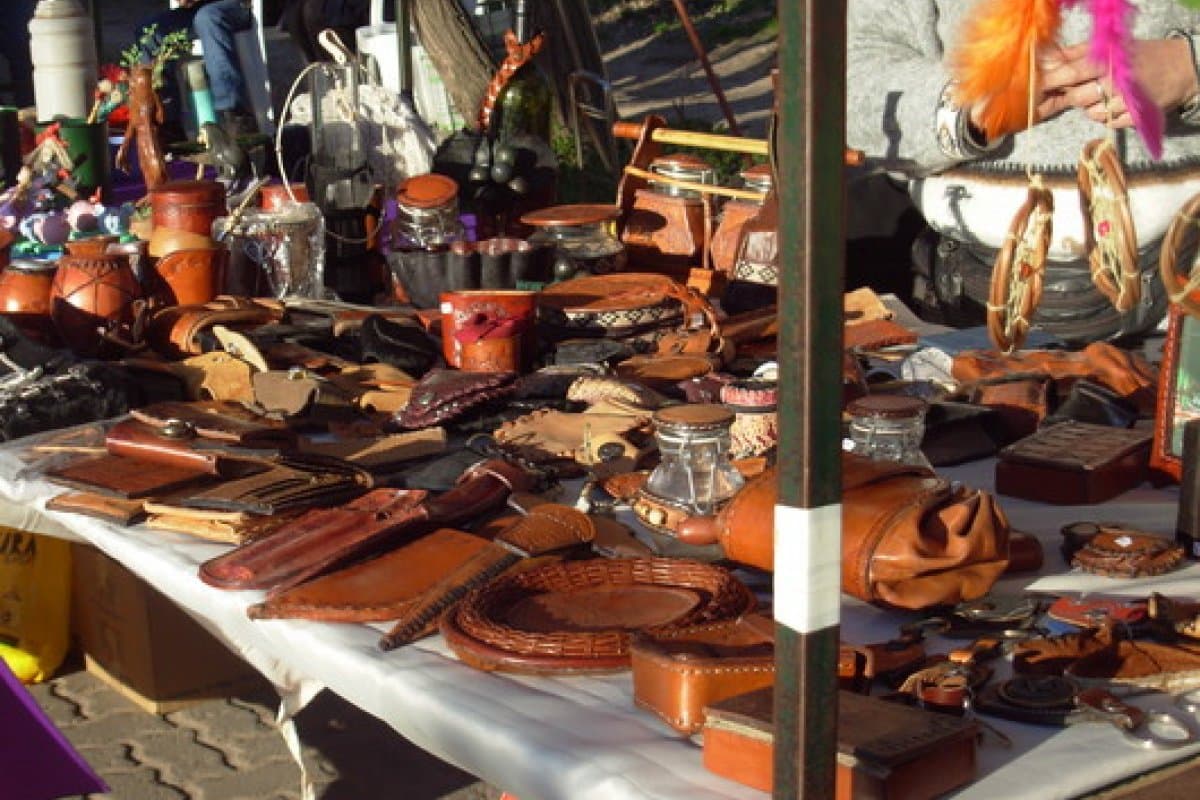 Mesa de un puesto de la feria cubierta de piezas de cuero trabajado: vainas de cuchillo, fundas, cintos, bandejas trenzadas y mates forrados.