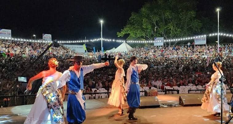 Parejas de folklore bailan de noche en un escenario al aire libre, frente a gradas colmadas de público.