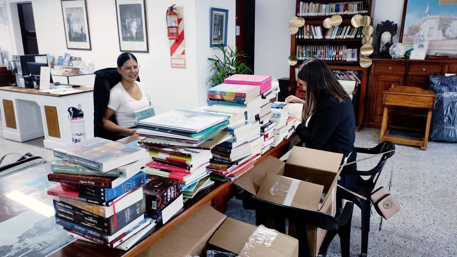 Sala de la Biblioteca Popular Rivadavia: dos mujeres ordenan pilas de libros sobre una mesa larga, con estantes y fotografías antiguas enmarcadas al fondo.