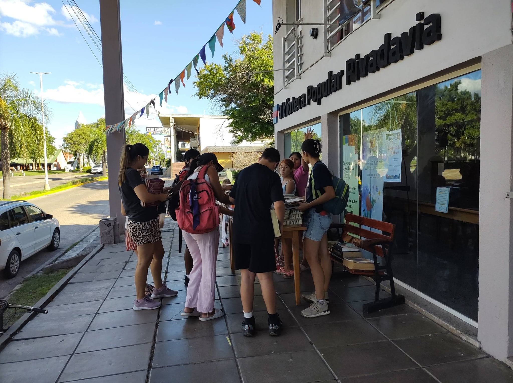 Vereda de la Biblioteca Popular Rivadavia: un grupo de personas elige libros de una mesa y un banco colocados frente a la vidriera.