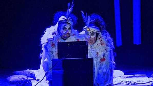 Dos intérpretes con maquillaje blanco y vestuario de plumas miran un viejo televisor en escena, bajo luz azul.